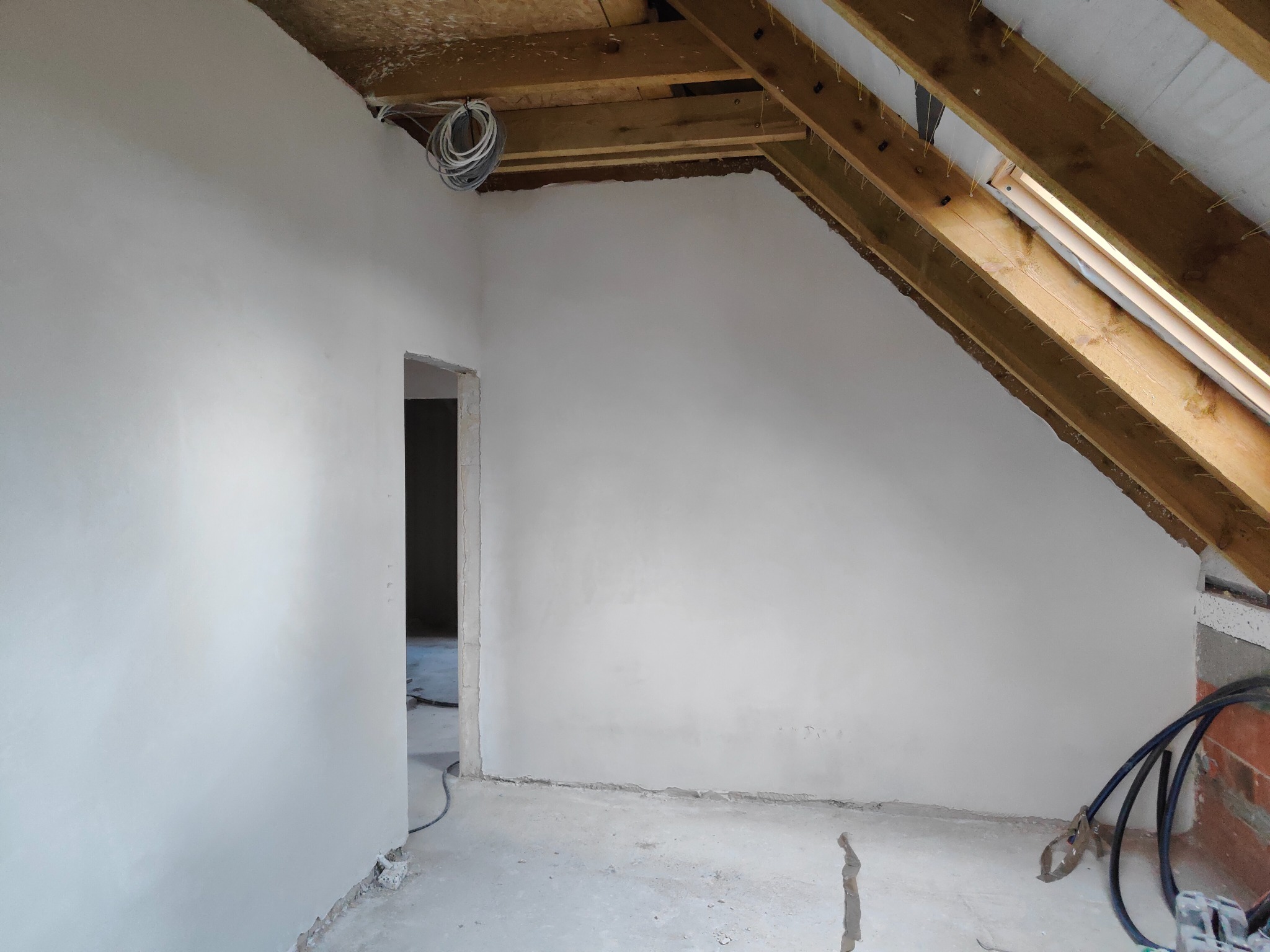 Świeżo otynkowane poddasze ze skośnym dachem, widoczne drewniane krokwie i wiązka przewodów elektrycznych zwisająca z sufitu.