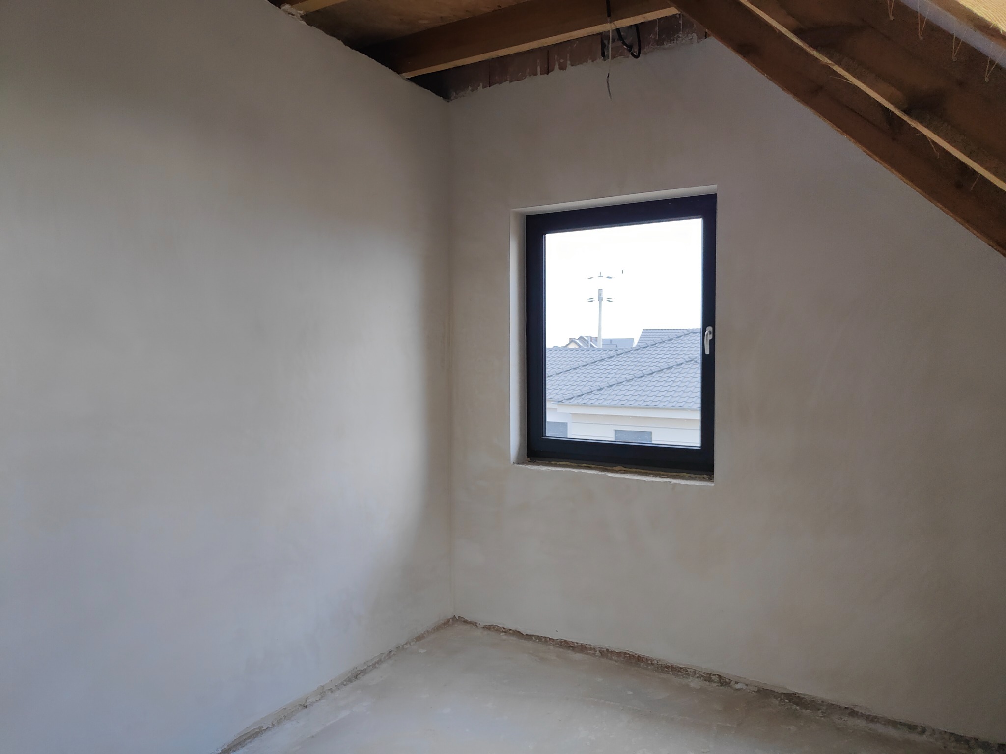 Pokój na poddaszu po nałożeniu gładzi gipsowej, widoczne surowe drewniane elementy konstrukcji dachu i czarne okno z widokiem na dach sąsiedniego domu.