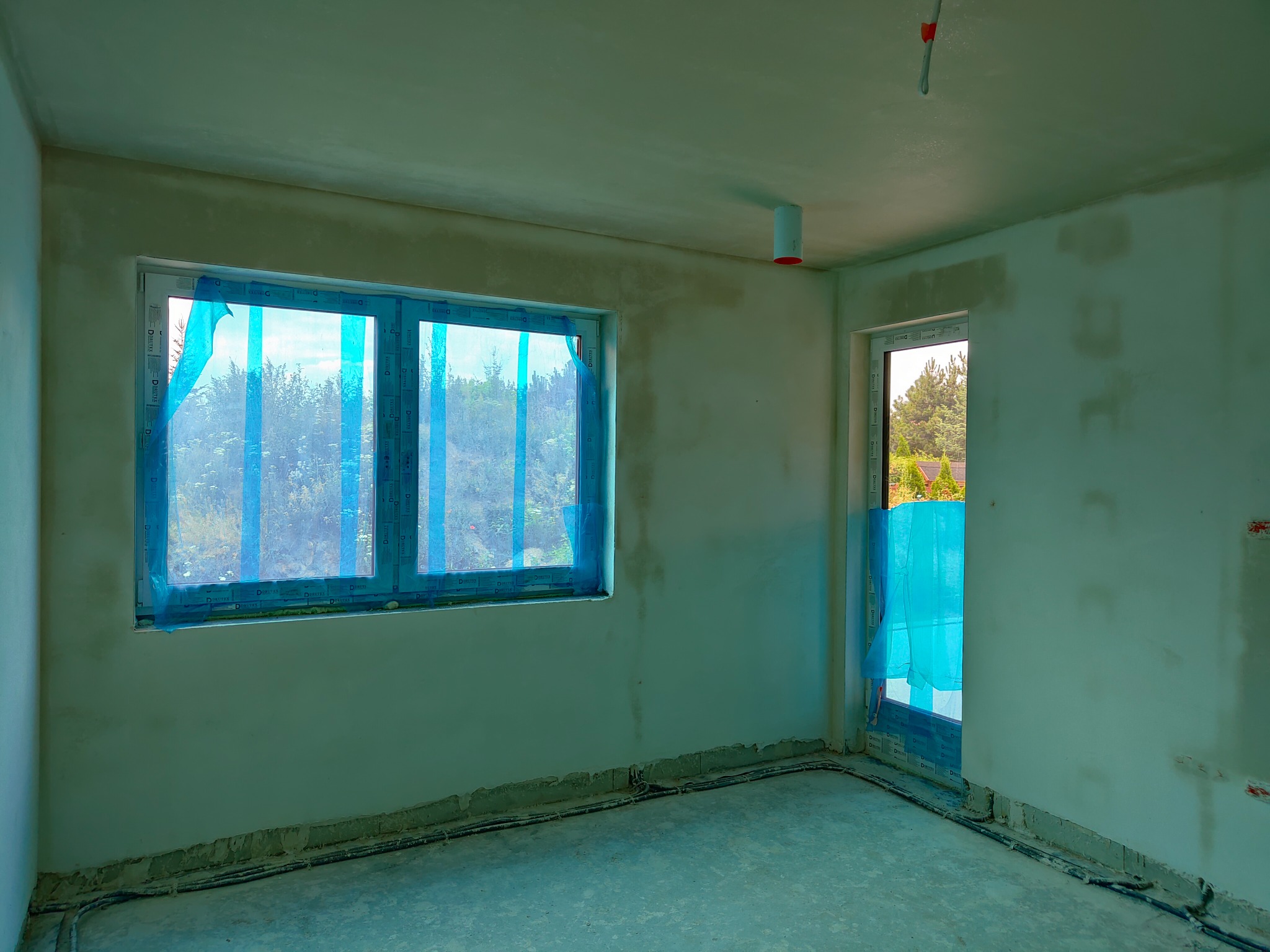 Wnętrze surowego pomieszczenia po nałożeniu tynku, z oknem i drzwiami zabezpieczonymi folią ochronną, widoczne przewody elektryczne przy podłodze.