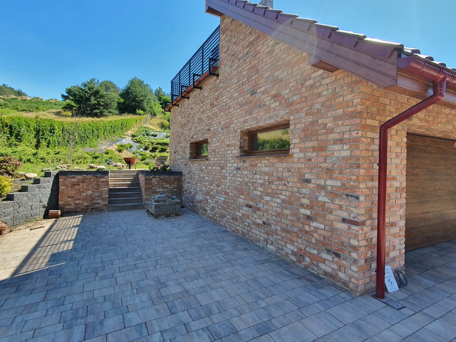 Nowoczesny dom z cegły klinkierowej z tarasem wyłożonym kostką brukową, widok na zagospodarowany ogród na wzgórzu.