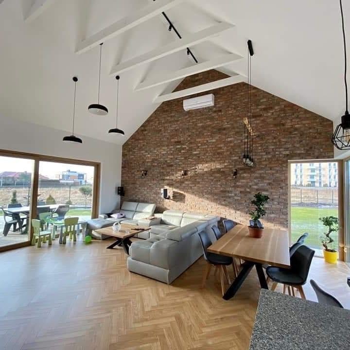 Przestronny salon z wysokim sufitem, drewnianą podłogą w jodełkę i ścianą z cegły, oświetlony wiszącymi lampami i naturalnym światłem wpadającym przez duże okno.