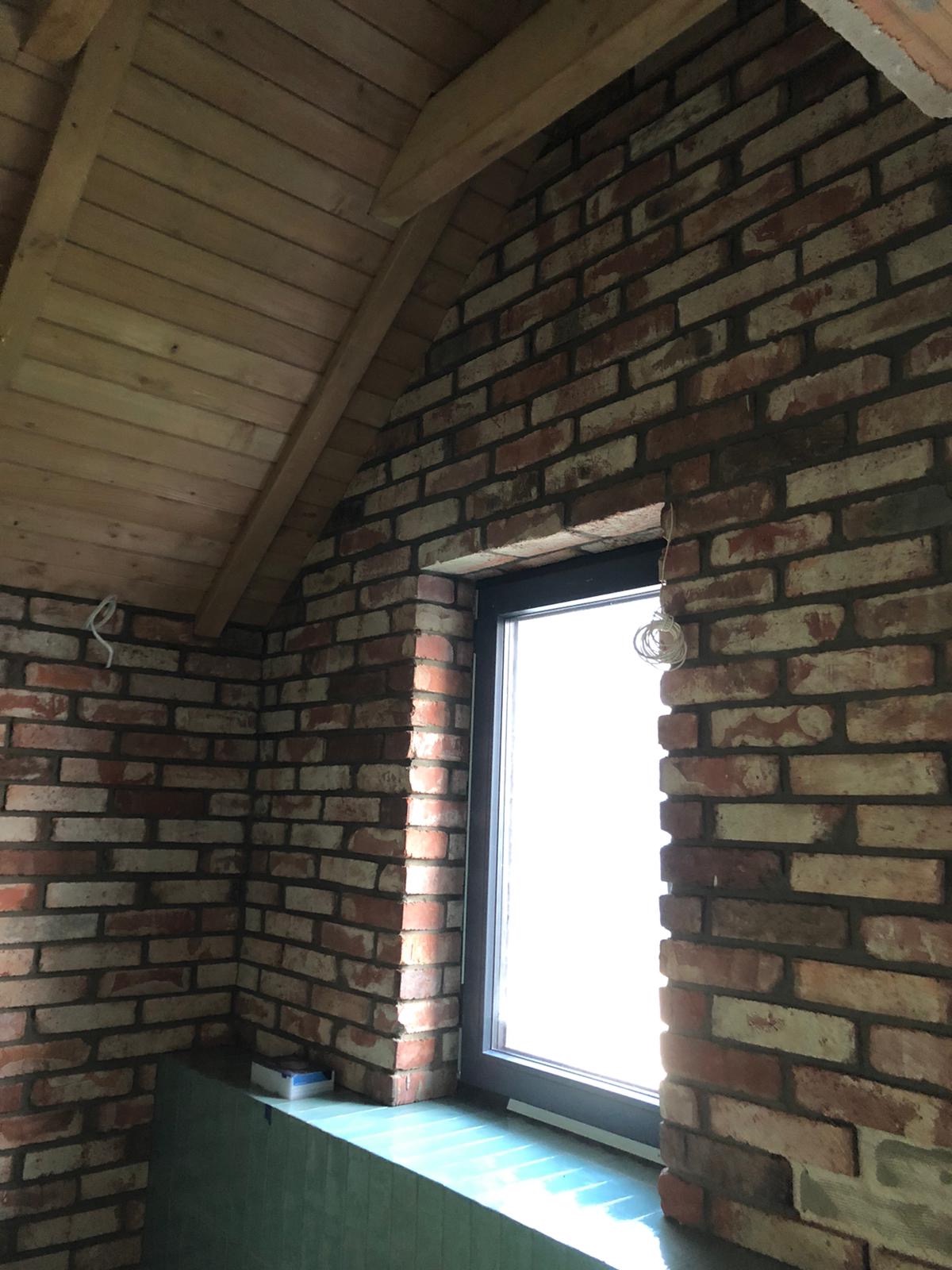 Wnętrze z surową czerwoną cegłą, drewnianym sufitem skośnym i oknem z czarną ramą, widoczne przewody elektryczne wystające ze ściany.