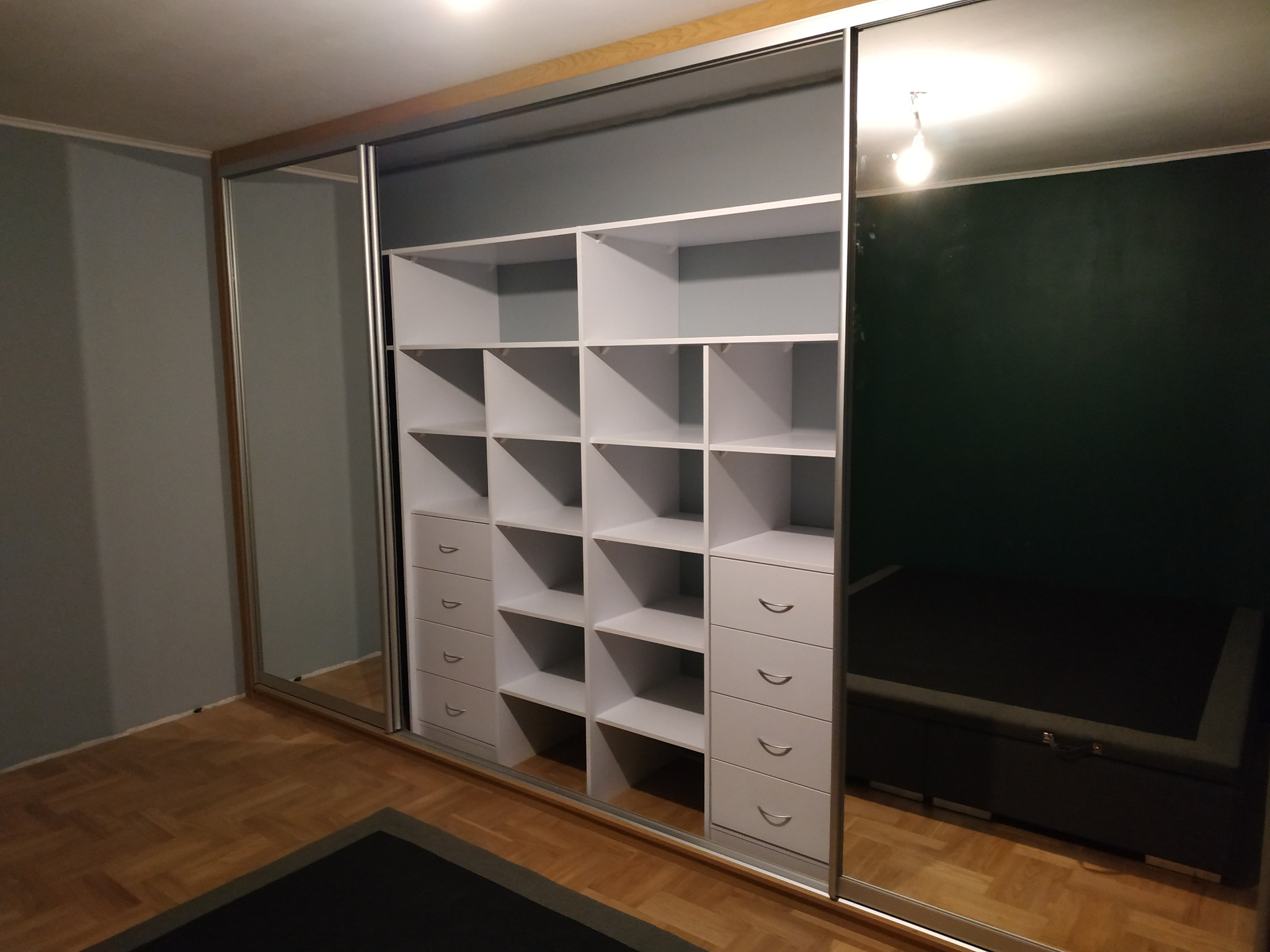 Biała szafa wnękowa z przesuwnymi lustrzanymi drzwiami, widoczne półki i szuflady, wnętrze w szarym odcieniu, oświetlenie sufitowe odbija się w lustrze.