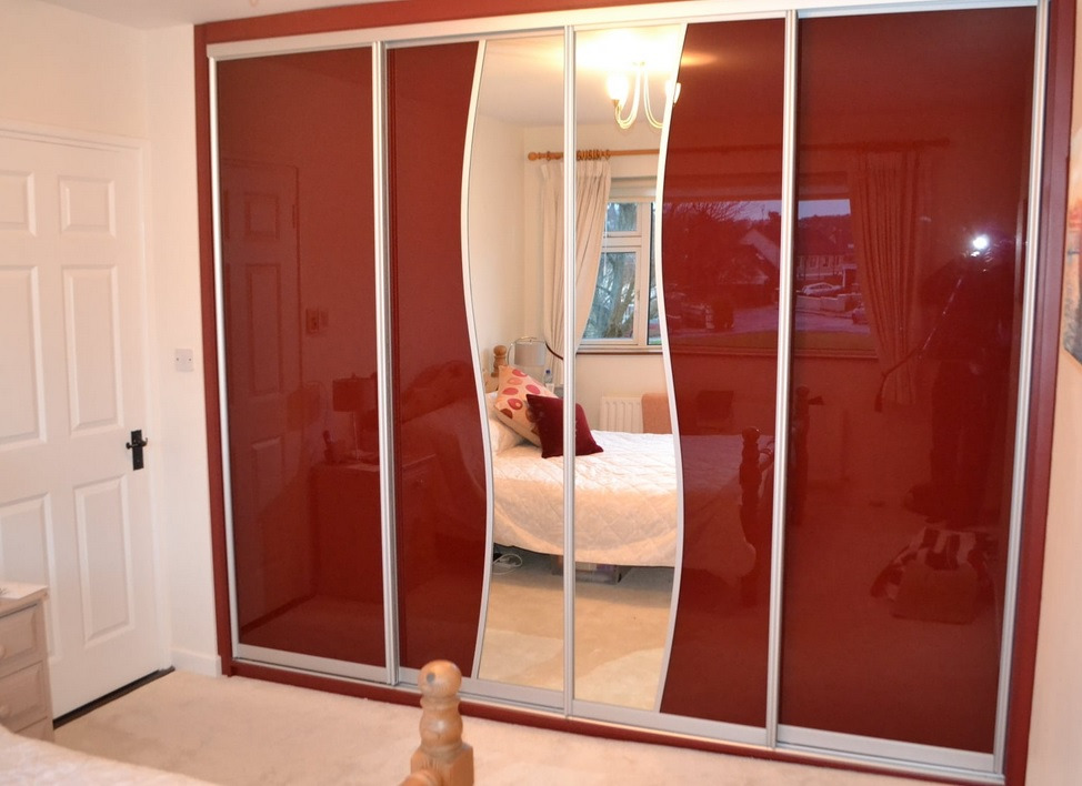 Szafa wnękowa z przesuwnymi drzwiami w kolorze bordowym z lustrzanymi panelami, odbijająca wnętrze sypialni z łóżkiem i lampą.