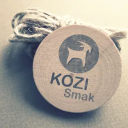 Okrągła drewniana zawieszka z logo firmy 'KOZI Smak' przedstawiająca stylizowaną kozę, przymocowana sznurkiem jutowym.
