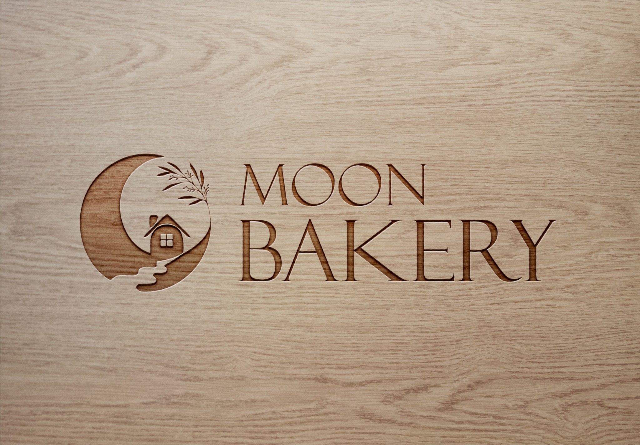 Logo 'Moon Bakery' wygrawerowane w jasnym drewnie, przedstawiające księżyc z domkiem, gałązką i napisem w klasycznym kroju pisma.