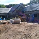 Budowa parterowego domu w trakcie: widoczne rusztowania, materiały budowlane na placu, grunt nierówny, w tle drzewa.