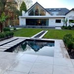 Nowoczesny dom z dużymi przeszkleniami i basenem w ogrodzie. Minimalistyczny design z białymi ścianami i ciemnym dachem. W tle zadbany trawnik i roślinność.