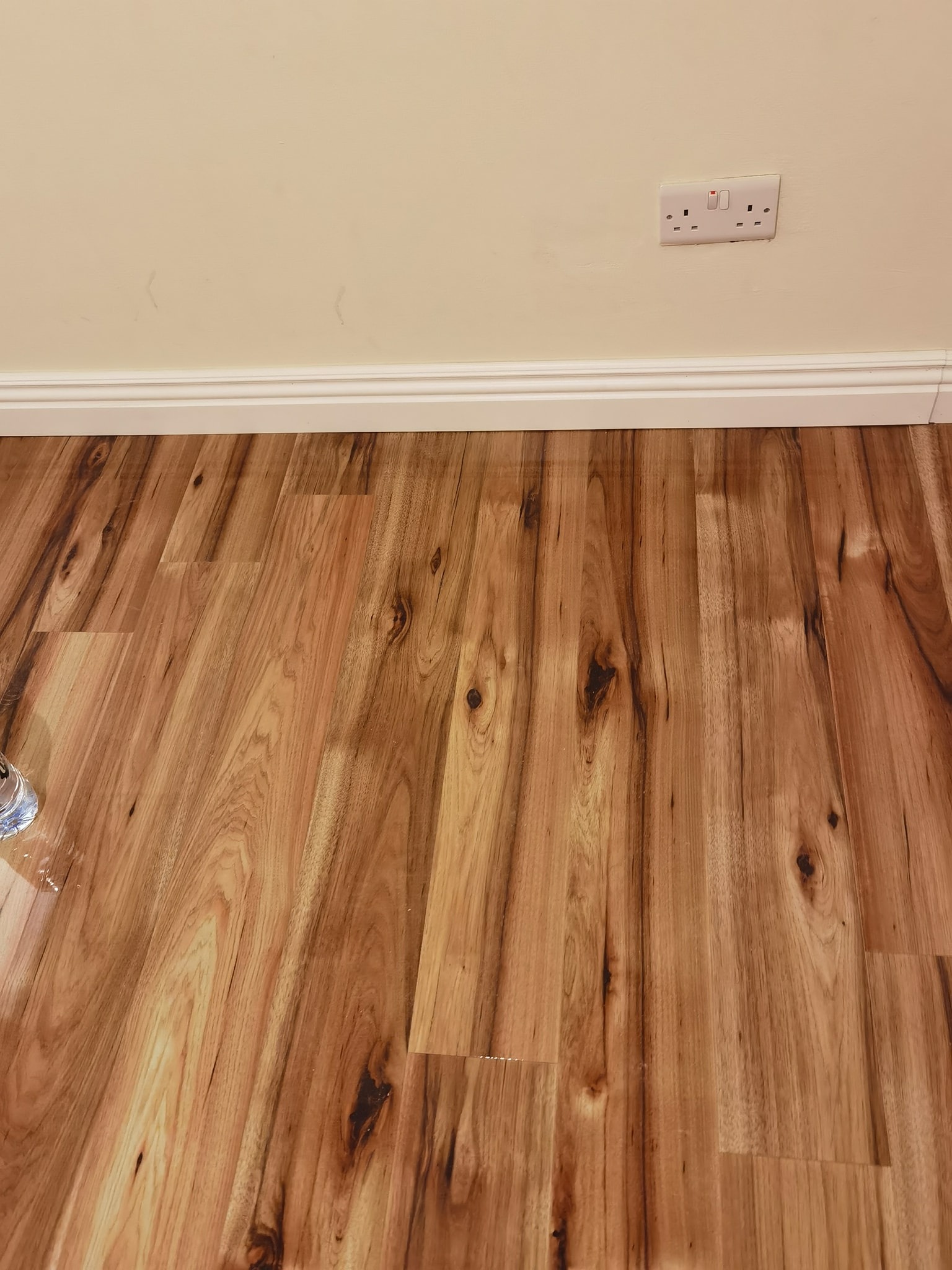 Ukośnie ułożona, drewnopodobna podłoga z widocznymi słojami i ciemnymi sękami, biała listwa przypodłogowa i kremowa ściana z gniazdkiem elektrycznym. W lewym dolnym rogu widoczny fragment szklanki.