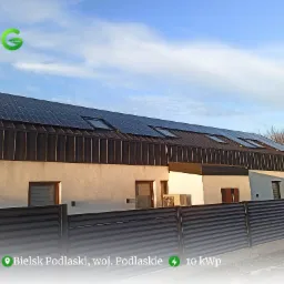 Dom jednorodzinny z zamontowanymi panelami słonecznymi na dachu z blachy trapezowej, widoczne okna dachowe i klimatyzator na ścianie, ogrodzenie panelowe w kolorze antracyt.