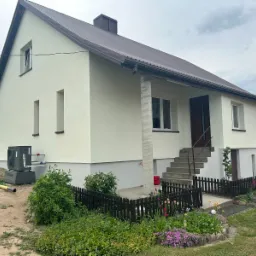 Dom jednorodzinny z jasną elewacją i brązowym dachem, na zewnątrz zainstalowana szara pompa ciepła z widocznymi rurami, ogródek z niskim płotkiem i kwiatami.