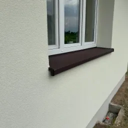 Elewacja budynku ocieplona styropianem z brązowym parapetem okiennym i widocznym fragmentem okna PCV