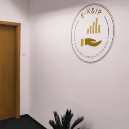 Wnętrze biura z logo firmy E-K&IP na ścianie, roślina doniczkowa w czarnej doniczce u dołu, widoczny fragment drzwi po lewej stronie.