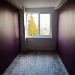 Pokój po malowaniu ścian w odcieniach fioletu i szarości, z położonymi panelami podłogowymi imitującymi drewno, widok na okno z drzewem na zewnątrz.