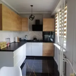 Nowoczesna kuchnia z czarnymi, błyszczącymi płytkami podłogowymi z żyłkami imitującymi marmur, białymi szafkami dolnymi i drewnopodobnymi szafkami górnymi, czarnym blatem oraz geometryczną lampą...