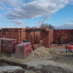 Wznoszenie ścian domu z czerwonej cegły Porotherm, widoczne palety cegieł, betoniarka, piasek, pracownik w tle i komin fabryczny w oddali pod błękitnym niebem z chmurami.