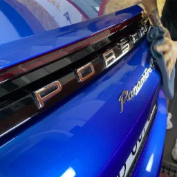 Błękitny samochód Porsche Panamera, tylna klapa czyszczona szarą ściereczką przez osobę z tatuażem na ramieniu, widoczne logo i tablica rejestracyjna.