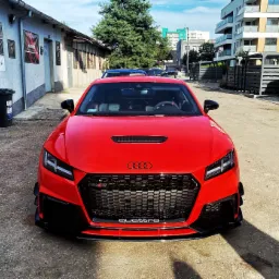 Czerwone Audi TT RS z przodu, zaparkowane na betonowym placu przed budynkiem warsztatowym z reklamami, w tle budynki mieszkalne.