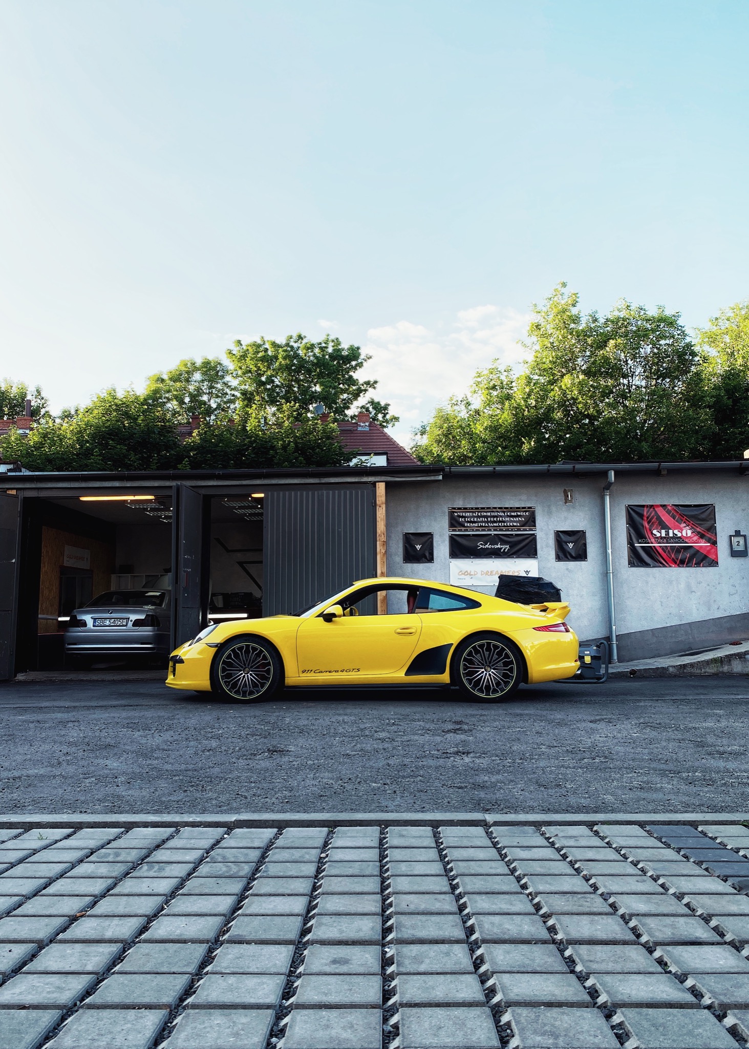 Żółte Porsche 911 Carrera 4 GTS zaparkowane przed garażem z szarym BMW w Zabrzu, widok z boku na tle budynku z reklamami.