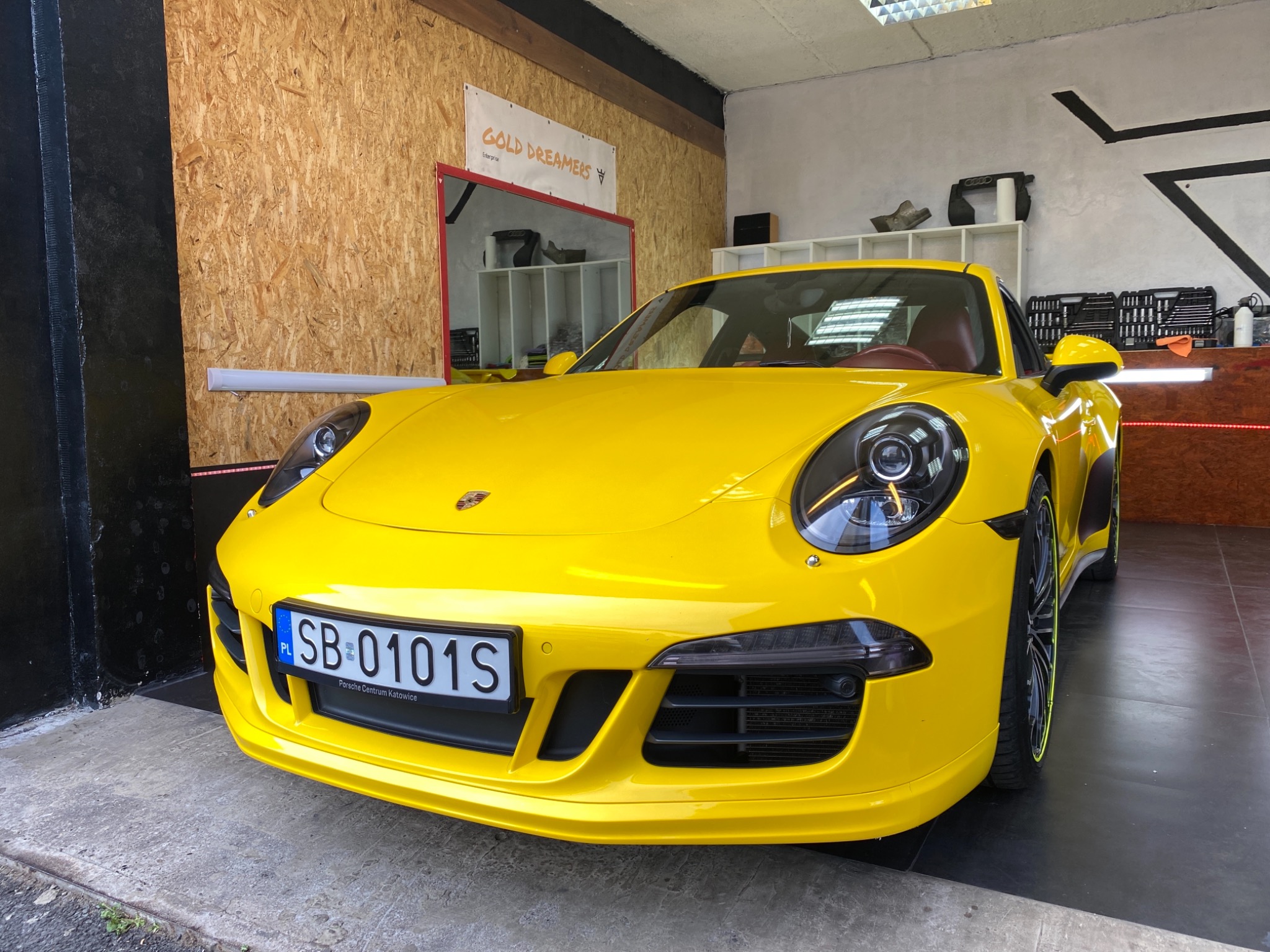 Żółte Porsche 911 w garażu z drewnianą ścianą i narzędziami w tle, widoczne tablice rejestracyjne SB 0101S.