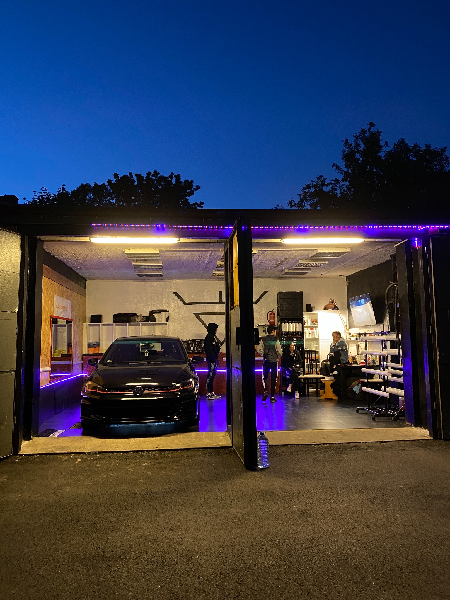 Czarny samochód w garażu z otwartymi drzwiami, oświetlony fioletowymi lampami LED, w tle widoczni ludzie.