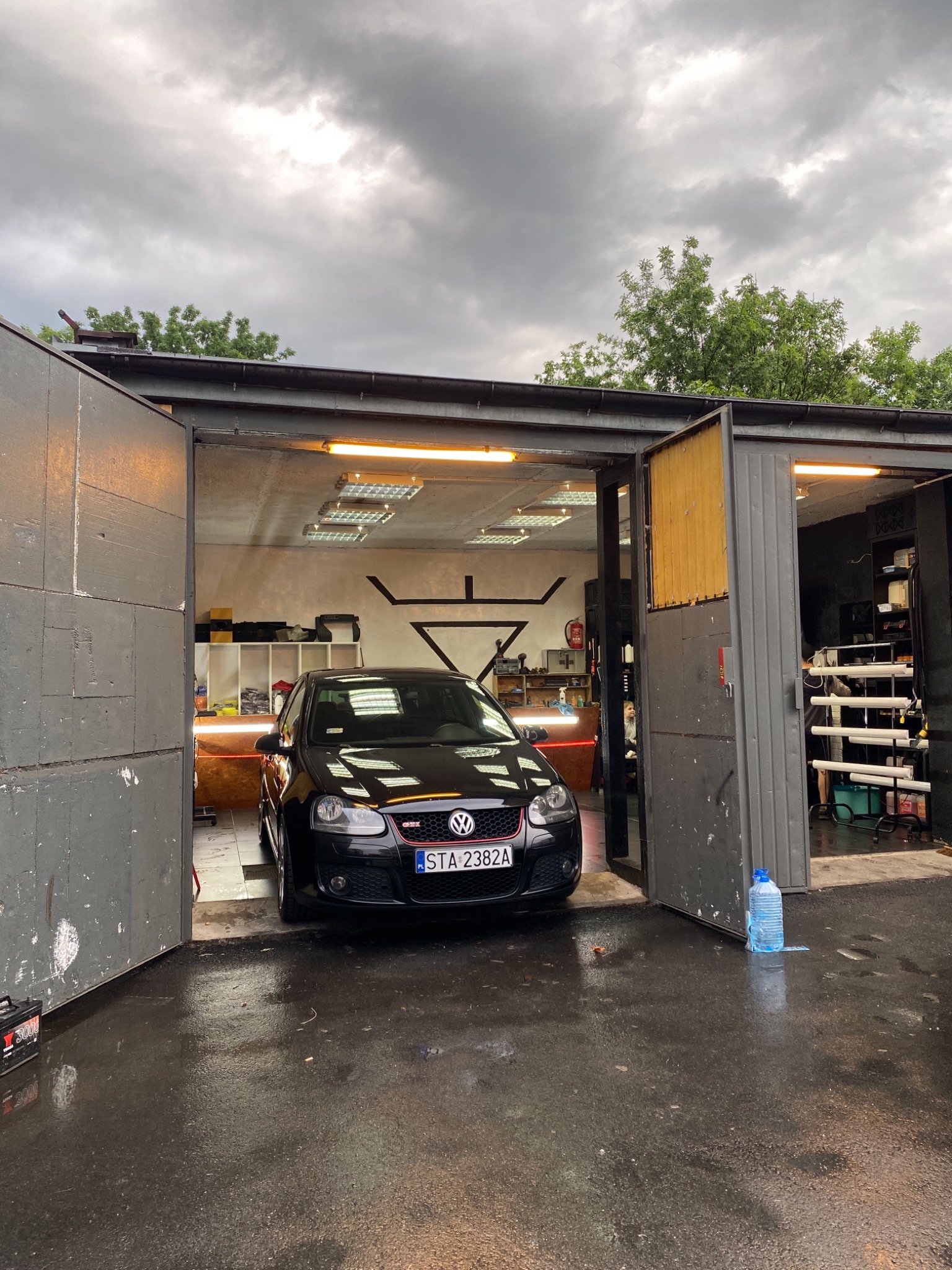 Czarny Volkswagen Golf GTI w warsztacie z otwartymi, szarymi drzwiami garażowymi, widoczne oświetlenie LED wewnątrz i mokra nawierzchnia przed garażem po deszczu.
