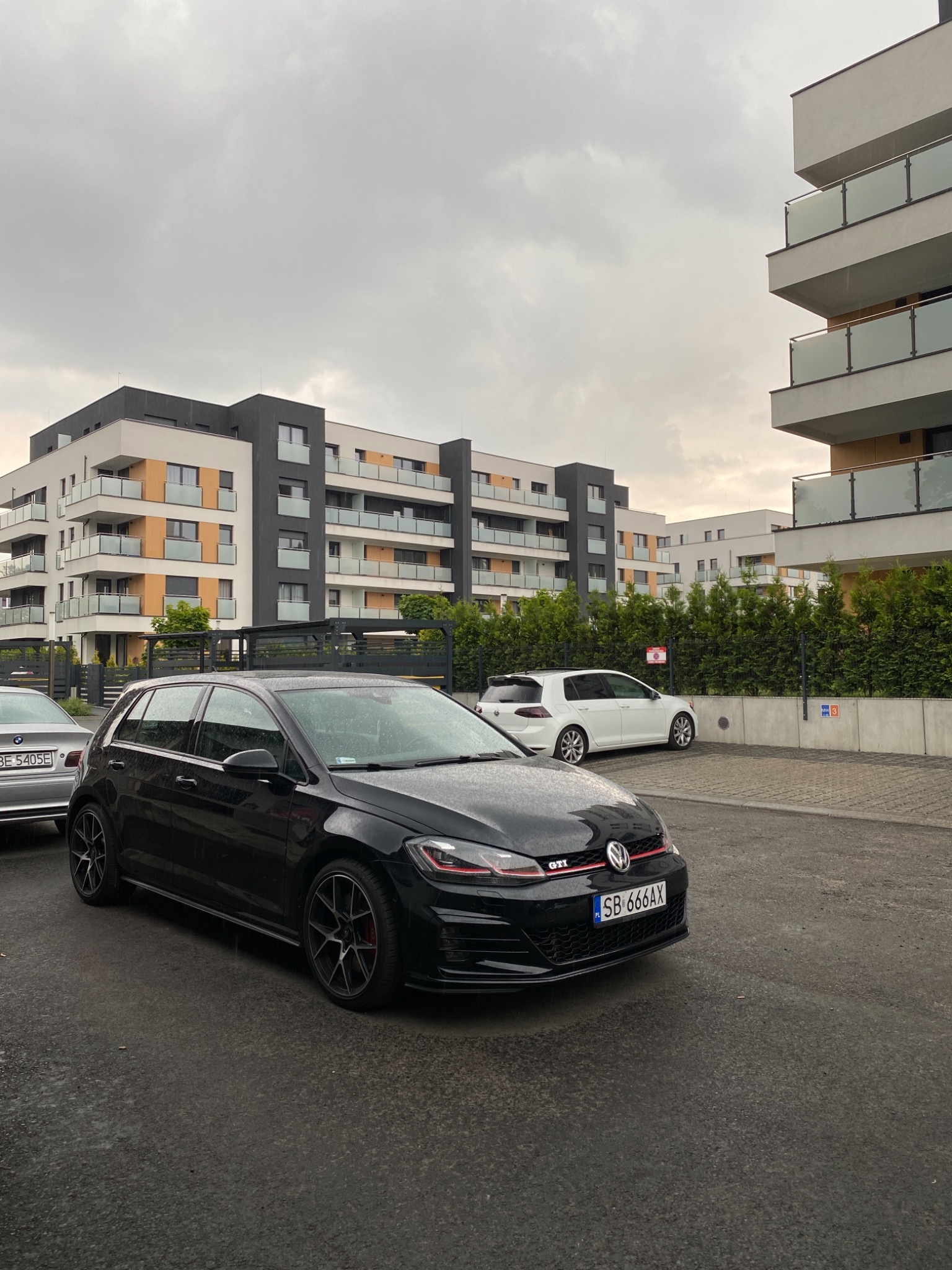 Czarny Volkswagen Golf GTI zaparkowany na mokrym parkingu przed nowoczesnym budynkiem mieszkalnym w pochmurny dzień. Widoczne krople wody na karoserii.