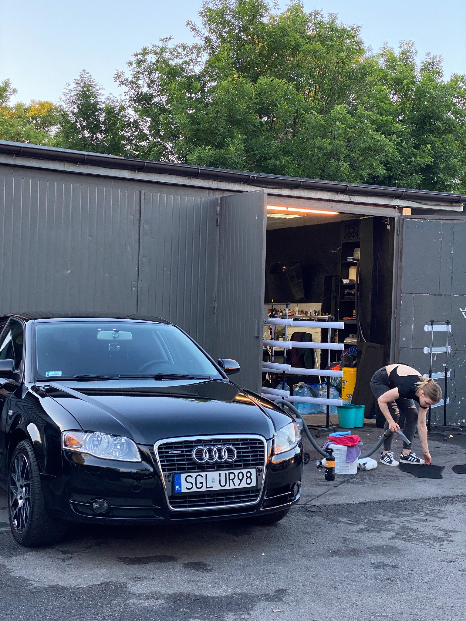 Czarny samochód Audi przed garażem; kobieta czyści dywanik samochodowy odkurzaczem; widoczne narzędzia i akcesoria do czyszczenia, butelki z płynami, wiadro z ściereczkami.