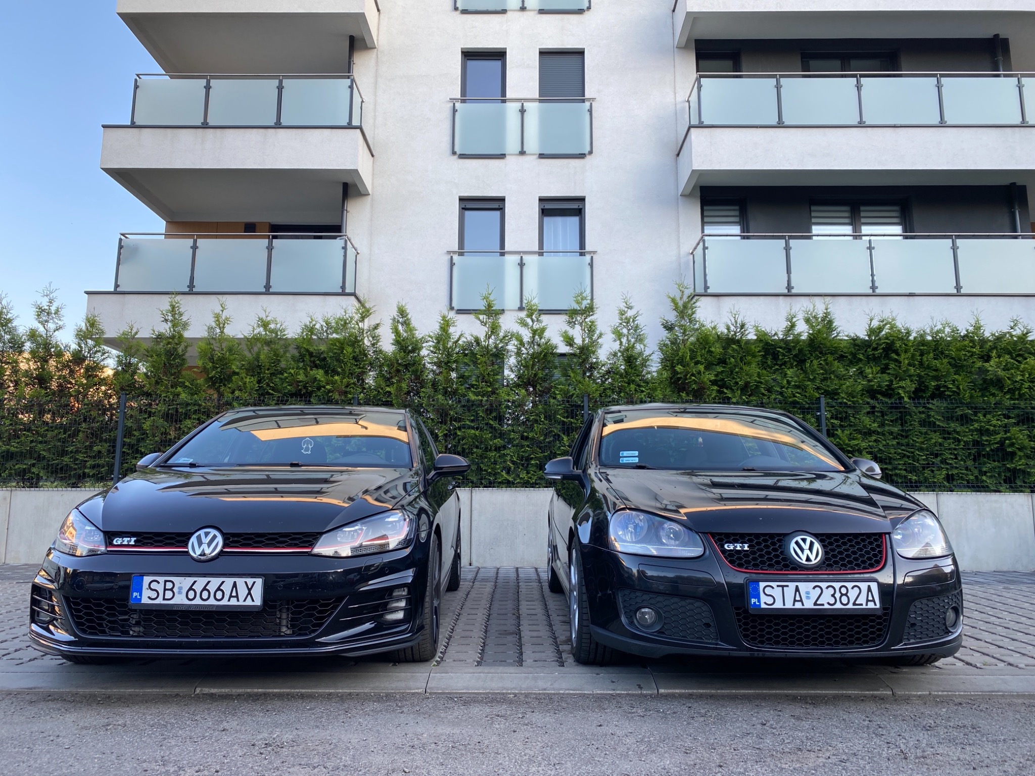 Dwa czarne samochody Volkswagen Golf GTI zaparkowane obok siebie na tle nowoczesnego budynku mieszkalnego z balkonami i zielonymi krzewami.