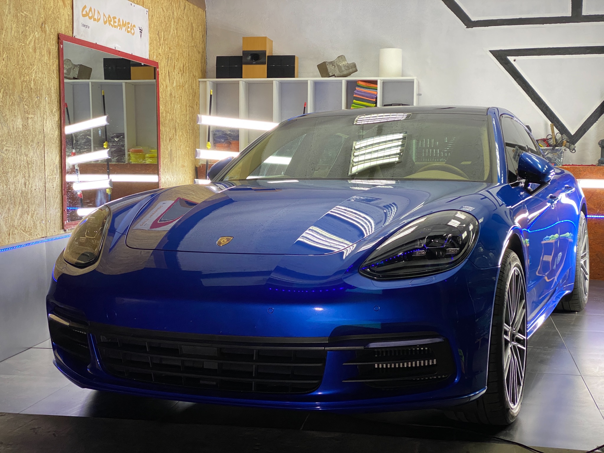 Błyszczący, niebieski Porsche Panamera po zabiegach pielęgnacyjnych, widoczne odbicia lamp w lakierze, w tle warsztat z półkami i akcesoriami.