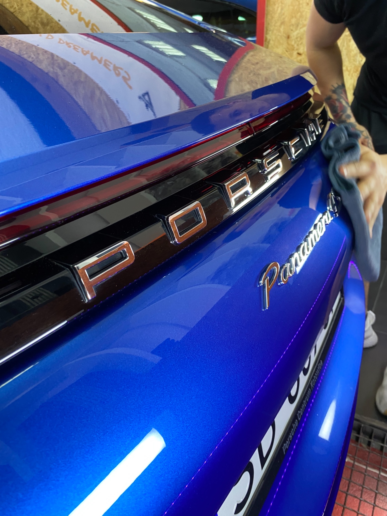 Błękitny samochód Porsche Panamera, tylna klapa czyszczona szarą ściereczką przez osobę z tatuażem na ramieniu, widoczne logo i tablica rejestracyjna.