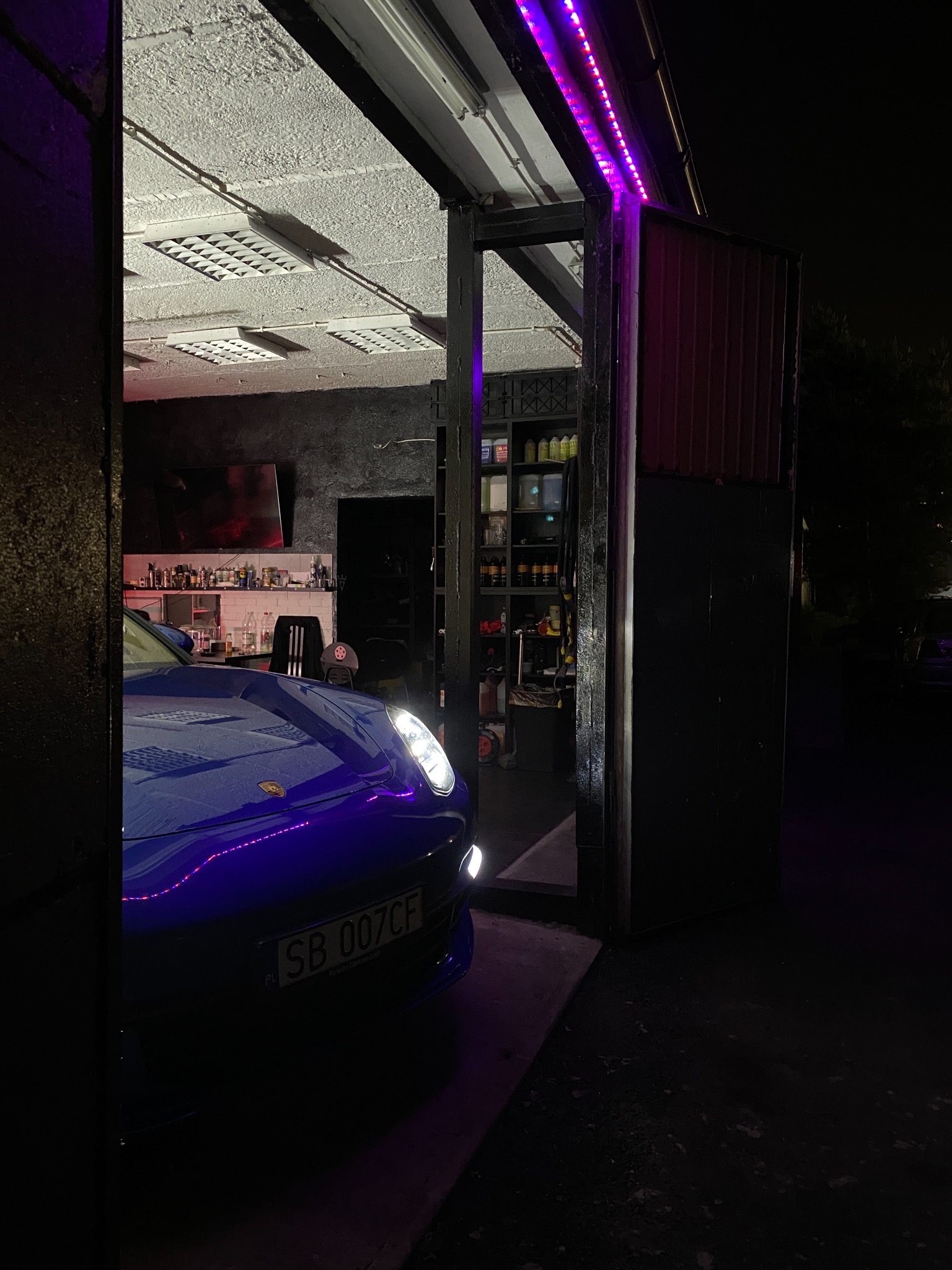 Niebieski samochód sportowy w garażu z oświetleniem LED, widok na półki z preparatami i narzędziami, ciemne tło.