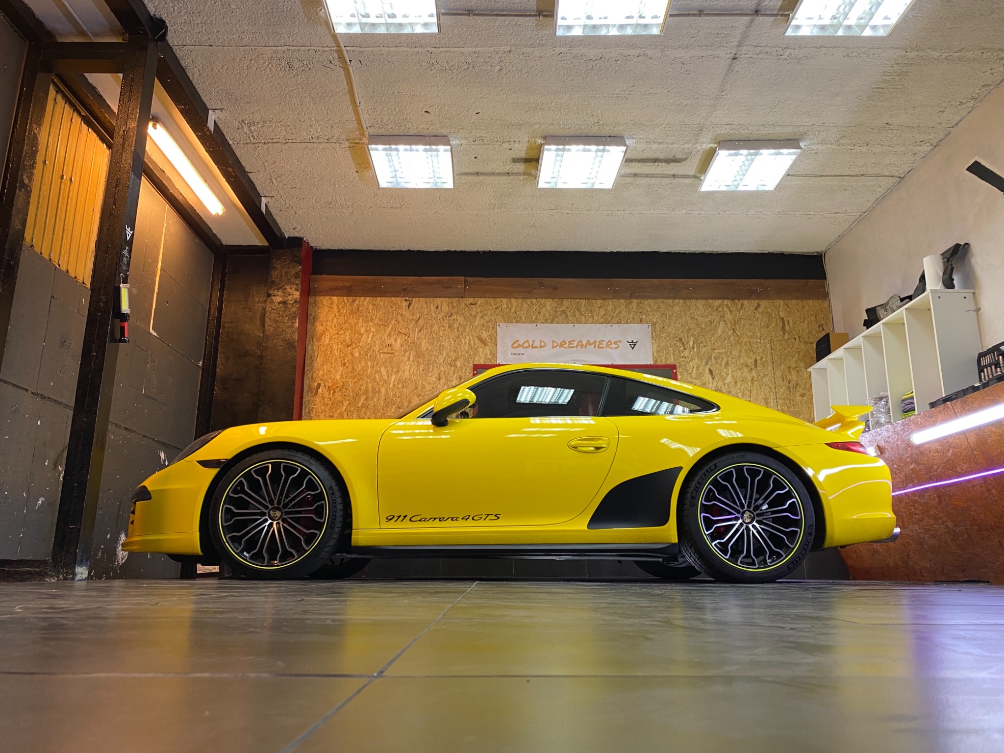 Żółty Porsche 911 Carrera 4 GTS, widok z boku w warsztacie, z widocznym logo Gold Dreamers w tle.