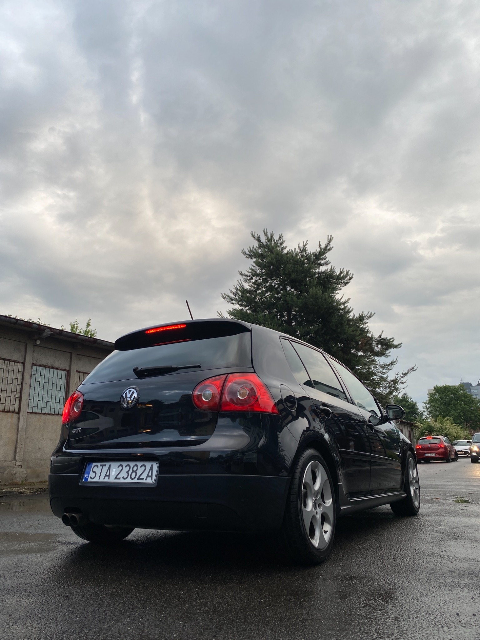 Czarny Volkswagen Golf GTI, widok z tyłu na mokrej nawierzchni, z widocznym budynkiem w tle i pochmurnym niebem.