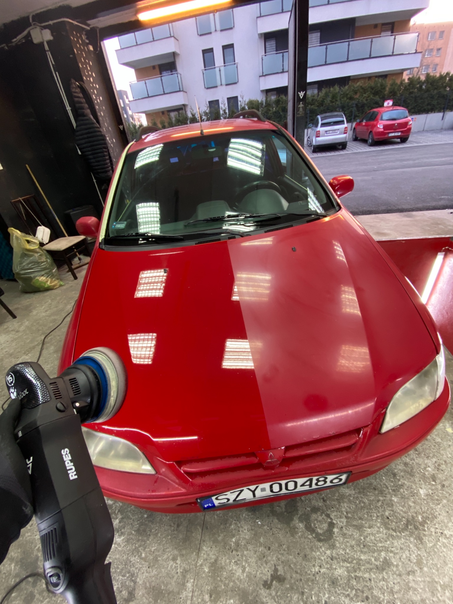 Czerwony samochód z widoczną różnicą w połysku lakieru na masce, polerowany za pomocą elektrycznej polerki Rupes. W tle budynek mieszkalny i inne samochody.