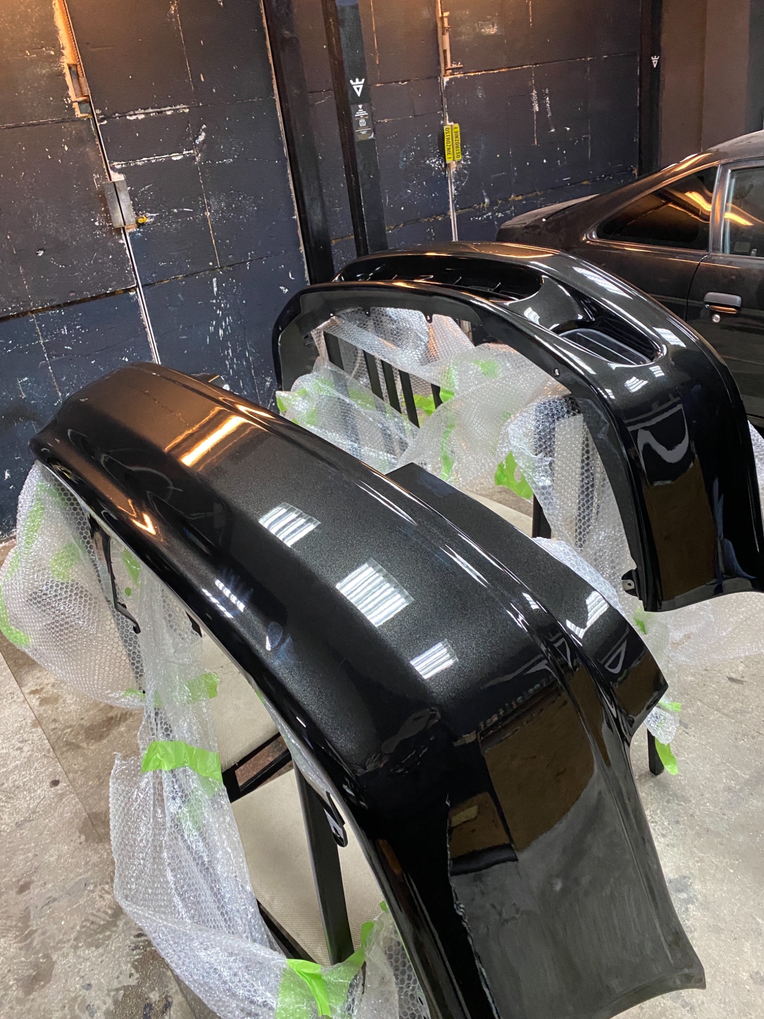 Dwa czarne, błyszczące zderzaki samochodowe, owinięte folią bąbelkową i zabezpieczone zieloną taśmą, stoją na podłodze warsztatu, w tle widoczny fragment samochodu i ciemne drzwi.