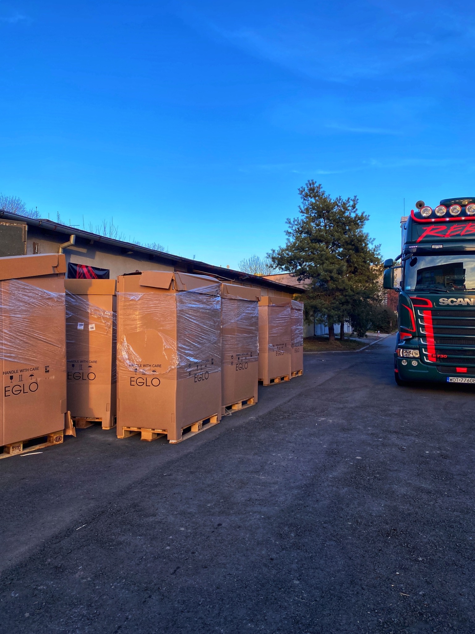 Paczki z logo EGLO na paletach obok zielonej ciężarówki Scania z napisem REBEL na tle błękitnego nieba i budynku.