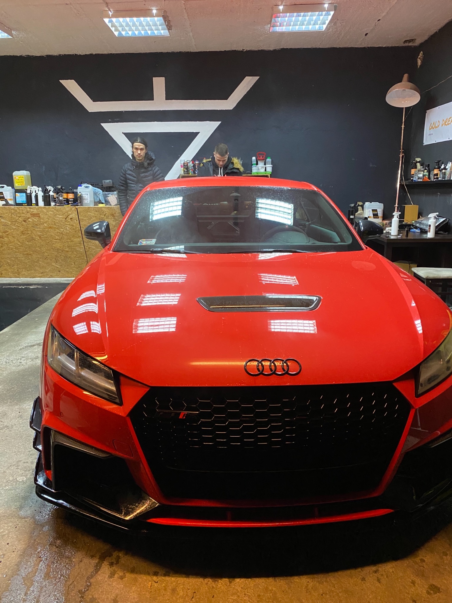 Czerwone Audi TT po umyciu, widoczne odbicia światła na lakierze, dwóch mężczyzn w tle, pomieszczenie z wyposażeniem do detailingu.