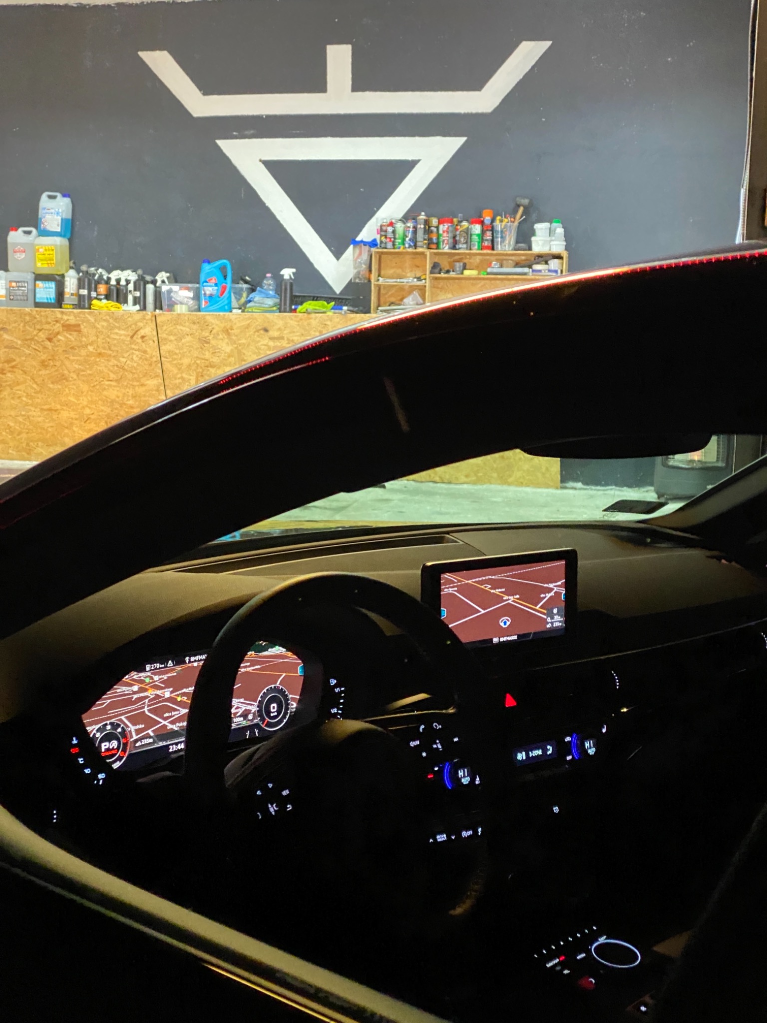 Wnętrze samochodu z włączonym ekranem nawigacji; w tle ściana warsztatu z logo i półkami pełnymi środków czyszczących.
