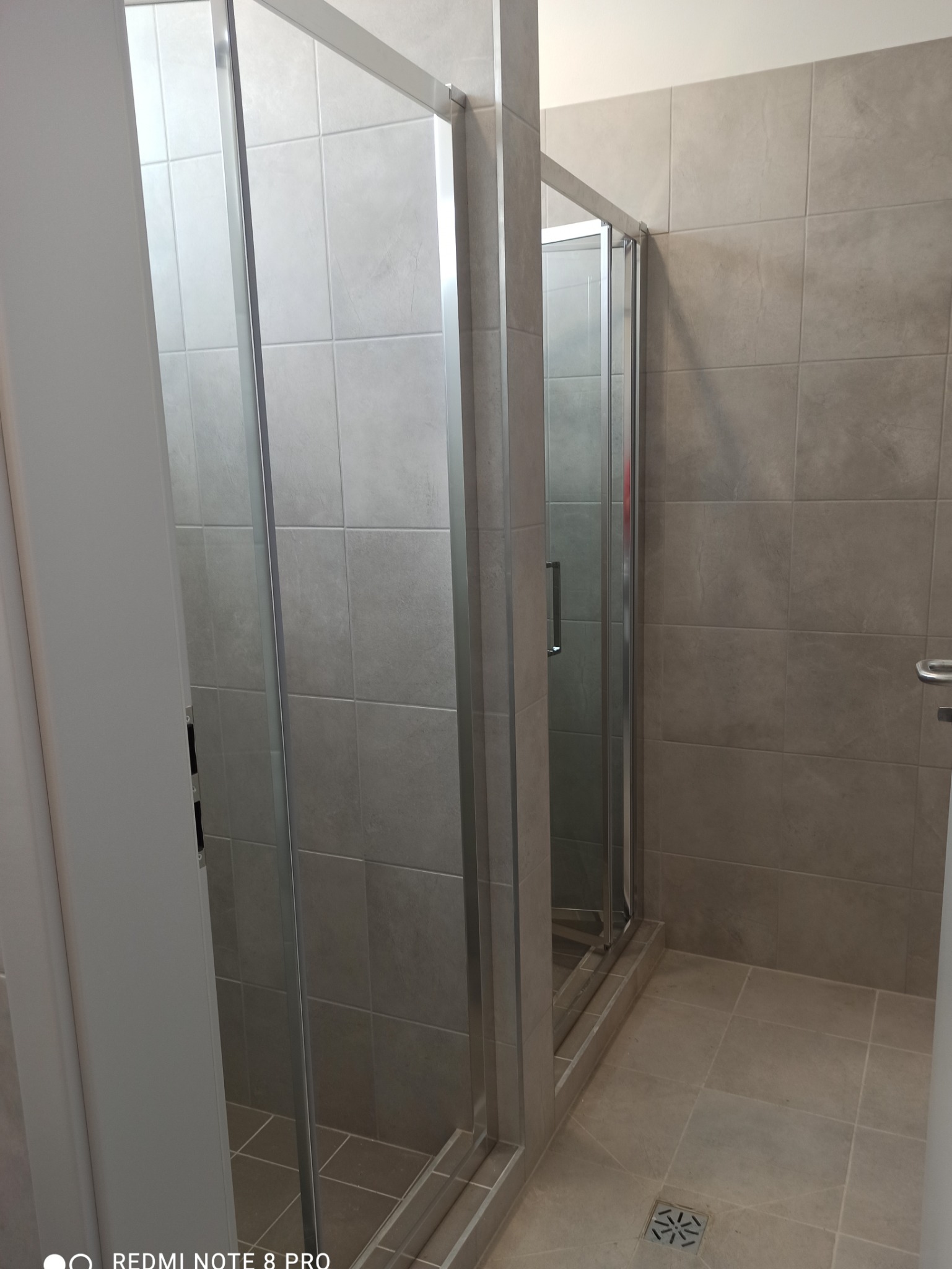 Szklana kabina prysznicowa z przesuwnymi drzwiami, wykończona szarymi, kwadratowymi płytkami ceramicznymi na ścianach i podłodze, z widocznym odpływem podłogowym.