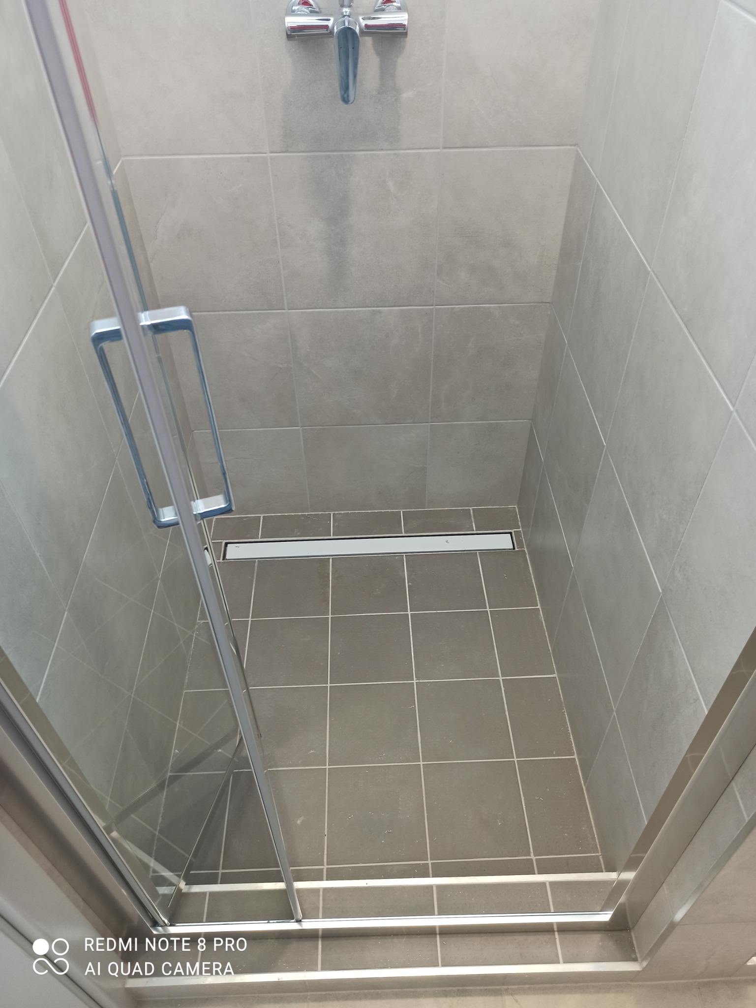 Wykończona kabina prysznicowa z szarymi płytkami, odpływ liniowy w podłodze, widoczna chromowana bateria i uchwyt szklanych drzwi.