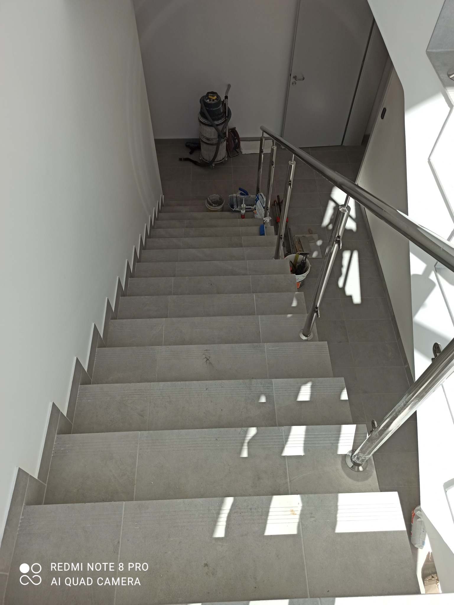 Wykończone schody wewnętrzne z szarych płytek z metalową balustradą, widok z góry, na górze widoczny odkurzacz przemysłowy i narzędzia.