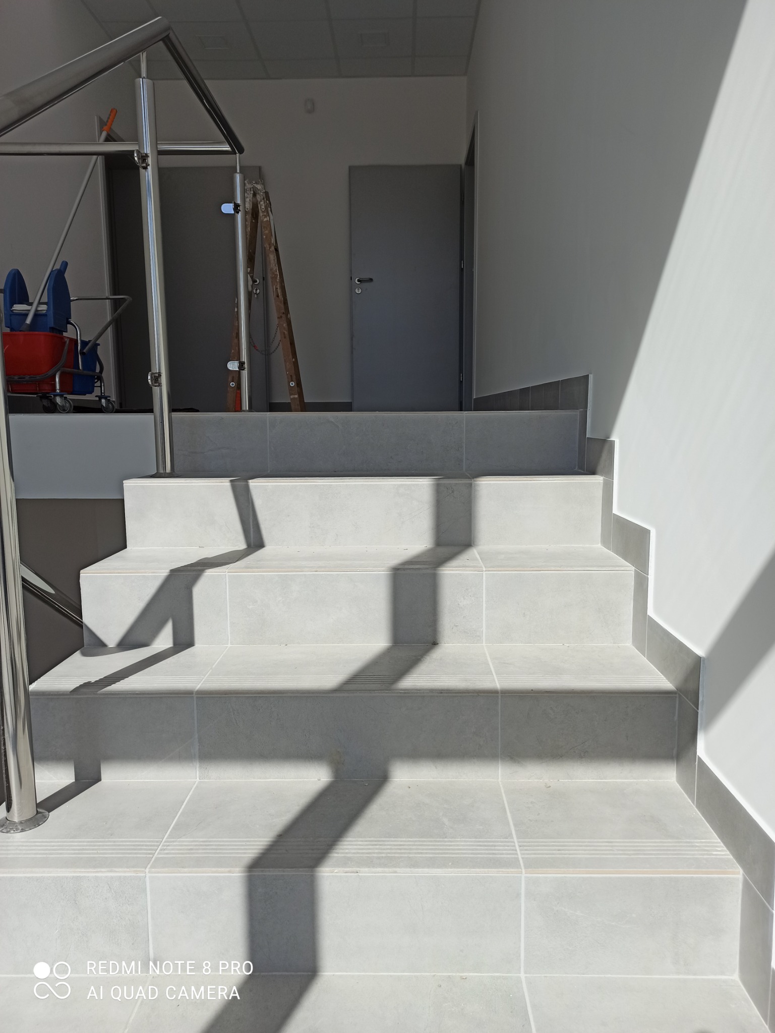Wykończone schody z szarych płytek ceramicznych z metalową barierką, widoczne w jasnym świetle słonecznym, z drabiną i wózkiem do sprzątania w tle.