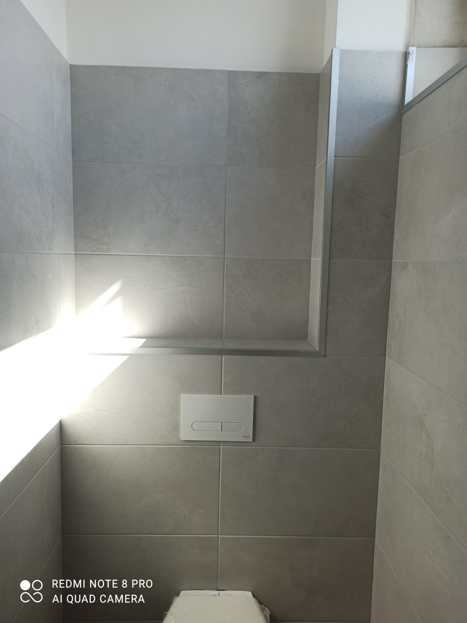 Szare, matowe płytki ceramiczne ułożone na ścianie w łazience, widoczny stelaż podtynkowy WC z przyciskiem spłukującym, fragment muszli klozetowej w folii ochronnej.