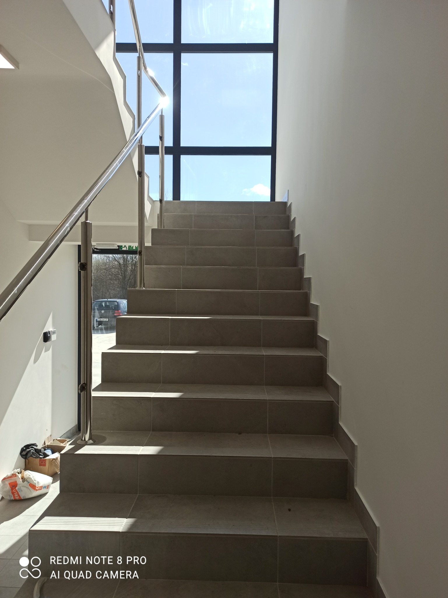 Schody wyłożone szarymi płytkami ceramicznymi z metalową barierką, widok z dołu, w tle okno z widokiem na błękitne niebo, na dole leżą kartony z materiałami budowlanymi.