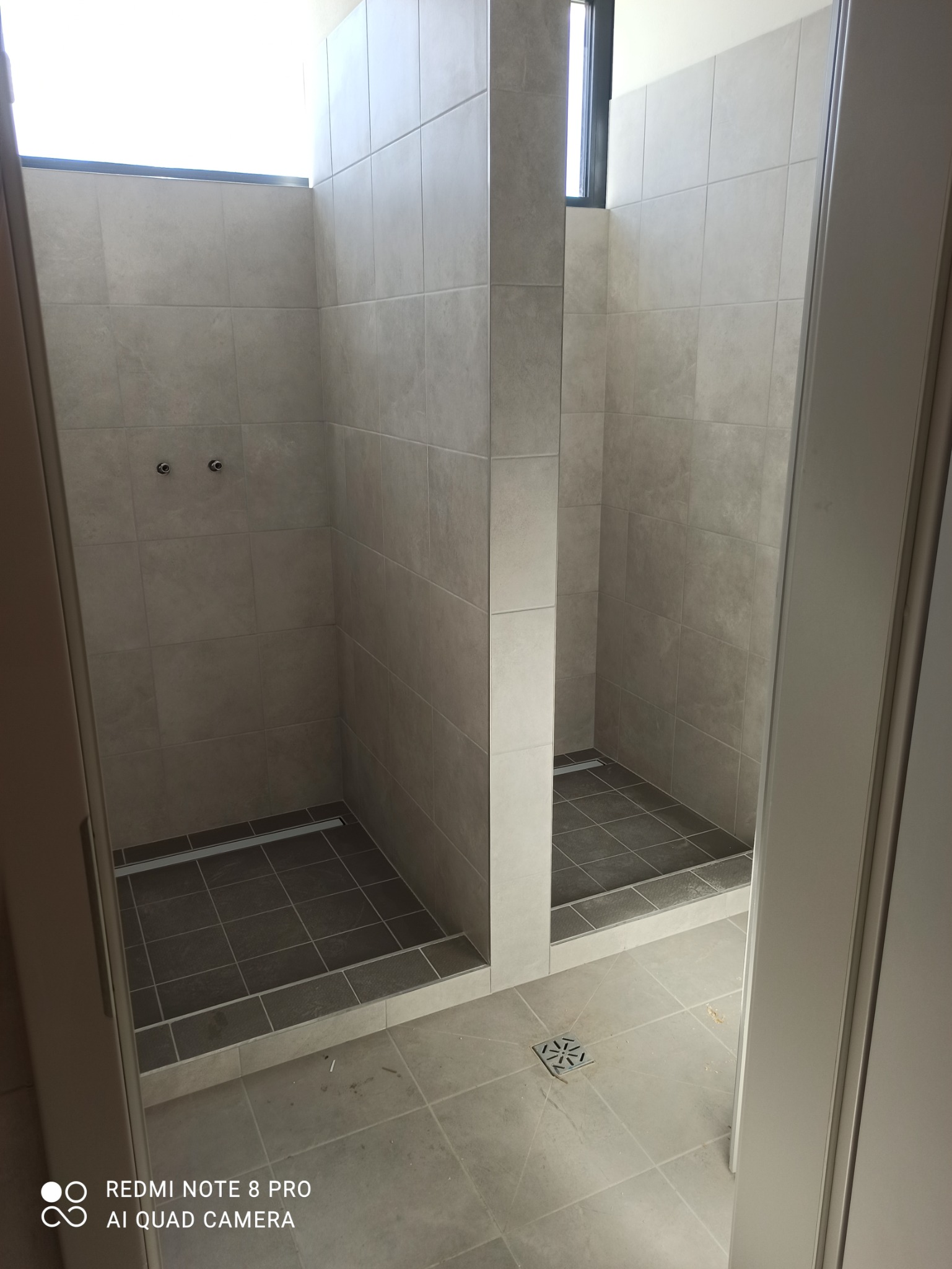 Dwa kabiny prysznicowe z jasnymi płytkami na ścianach i ciemnymi płytkami na podłodze, oddzielone ścianką, widoczne odpływy liniowe i punktowe.