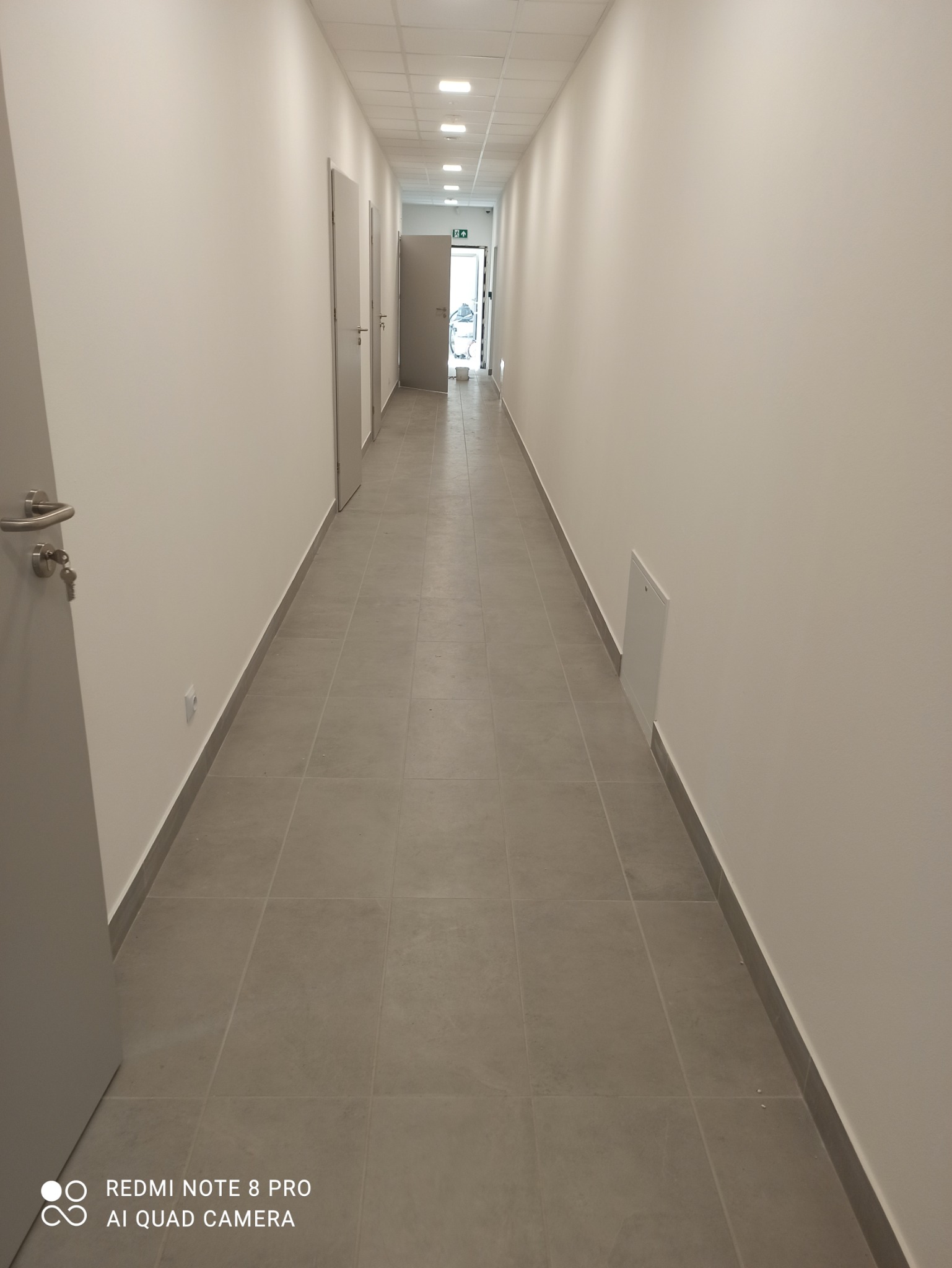 Długi, pusty korytarz z szarą podłogą wyłożoną płytkami, białymi ścianami i kilkoma drzwiami po obu stronach, oświetlony przez równomiernie rozmieszczone lampy sufitowe. Na pierwszym planie...