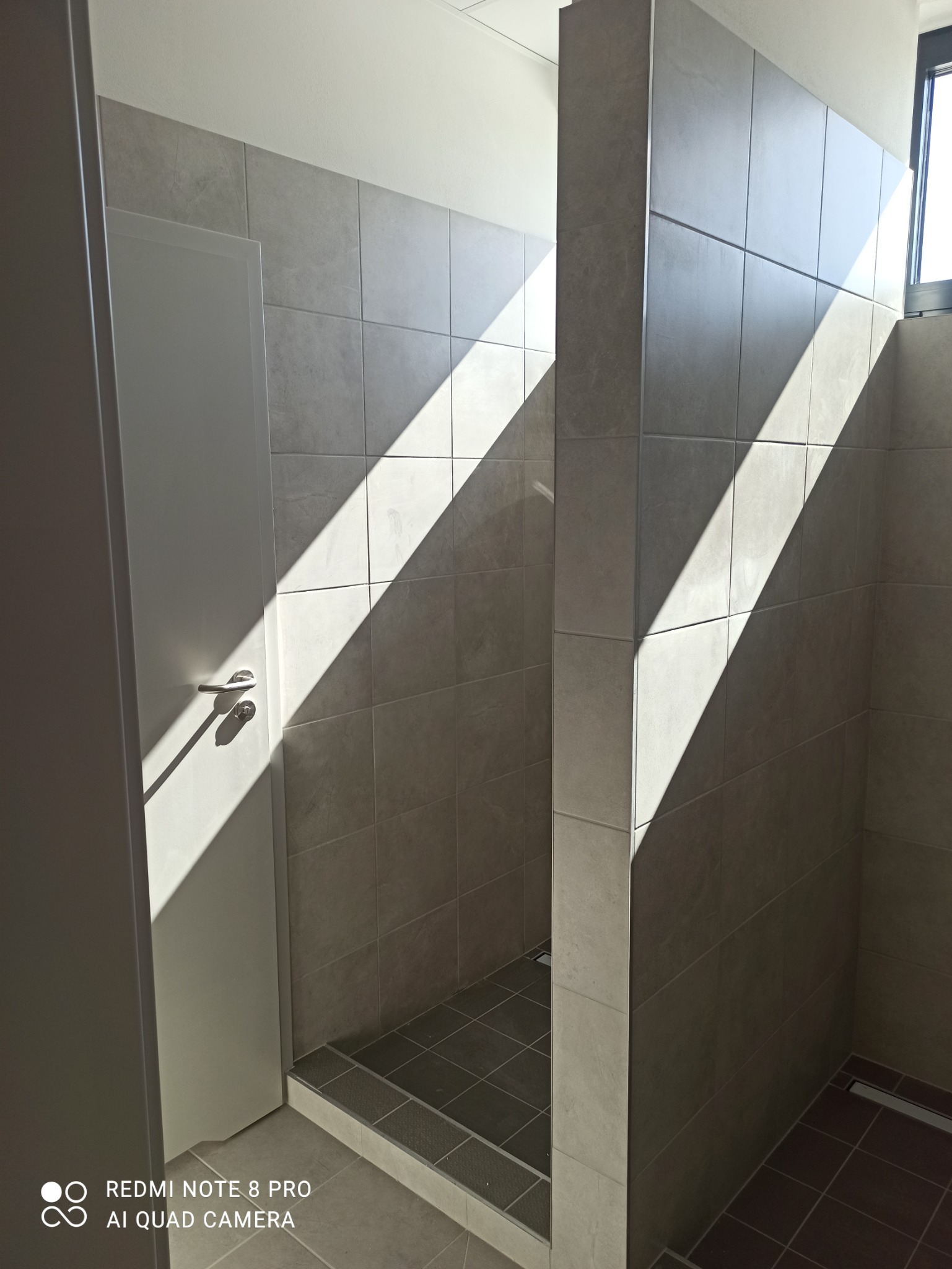 Wykończona kabina prysznicowa z szarymi płytkami, widoczne ostre światło słoneczne padające przez okno, podkreślające fakturę płytek. Zastosowano ciemniejsze płytki na podłodze brodzika.