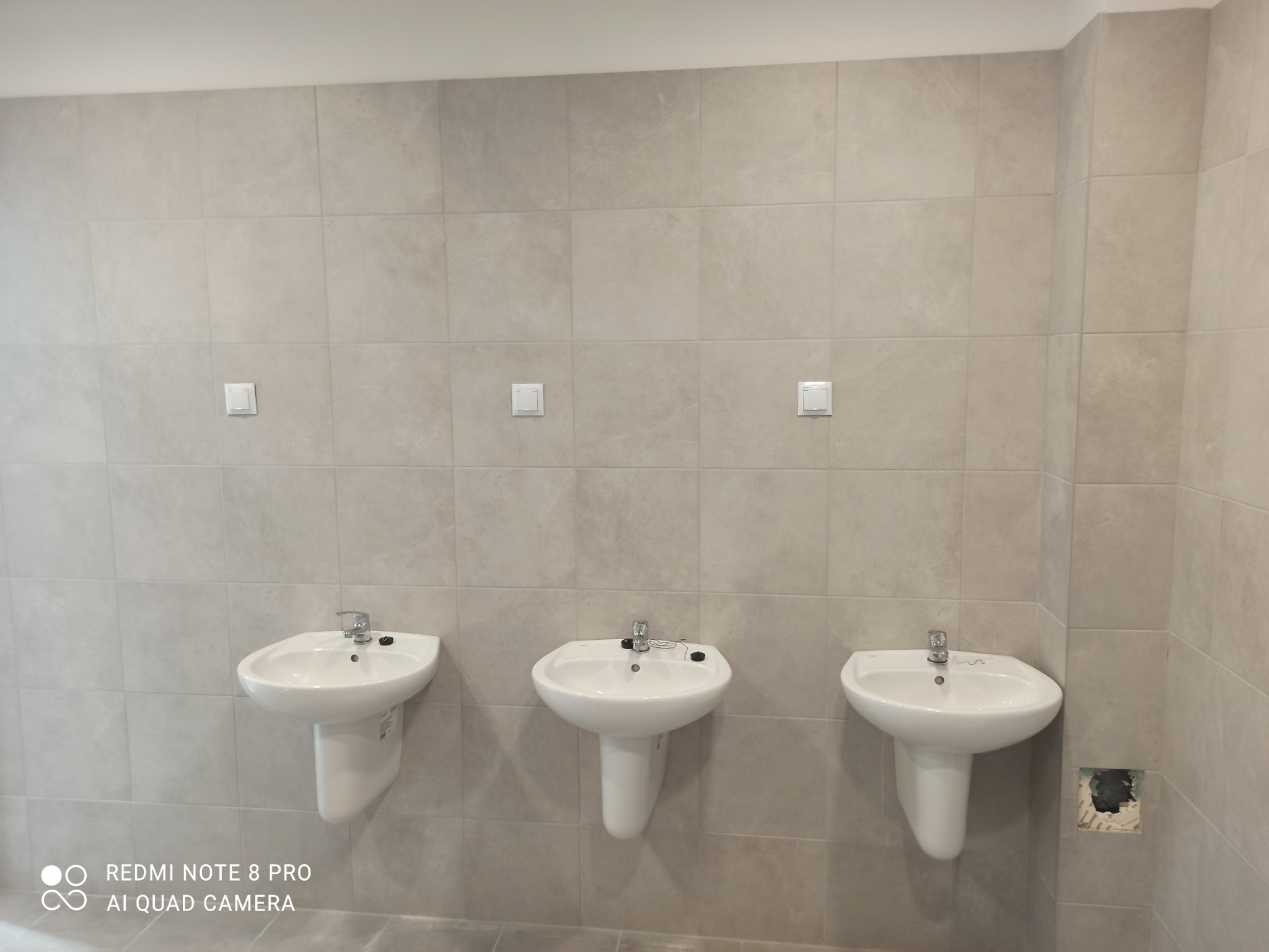 Wykończona łazienka publiczna z trzema białymi umywalkami ceramicznymi zamontowanymi na ścianie wyłożonej szarymi płytkami, z widocznymi włącznikami światła i niedokończonym otworem instalacyjnym...