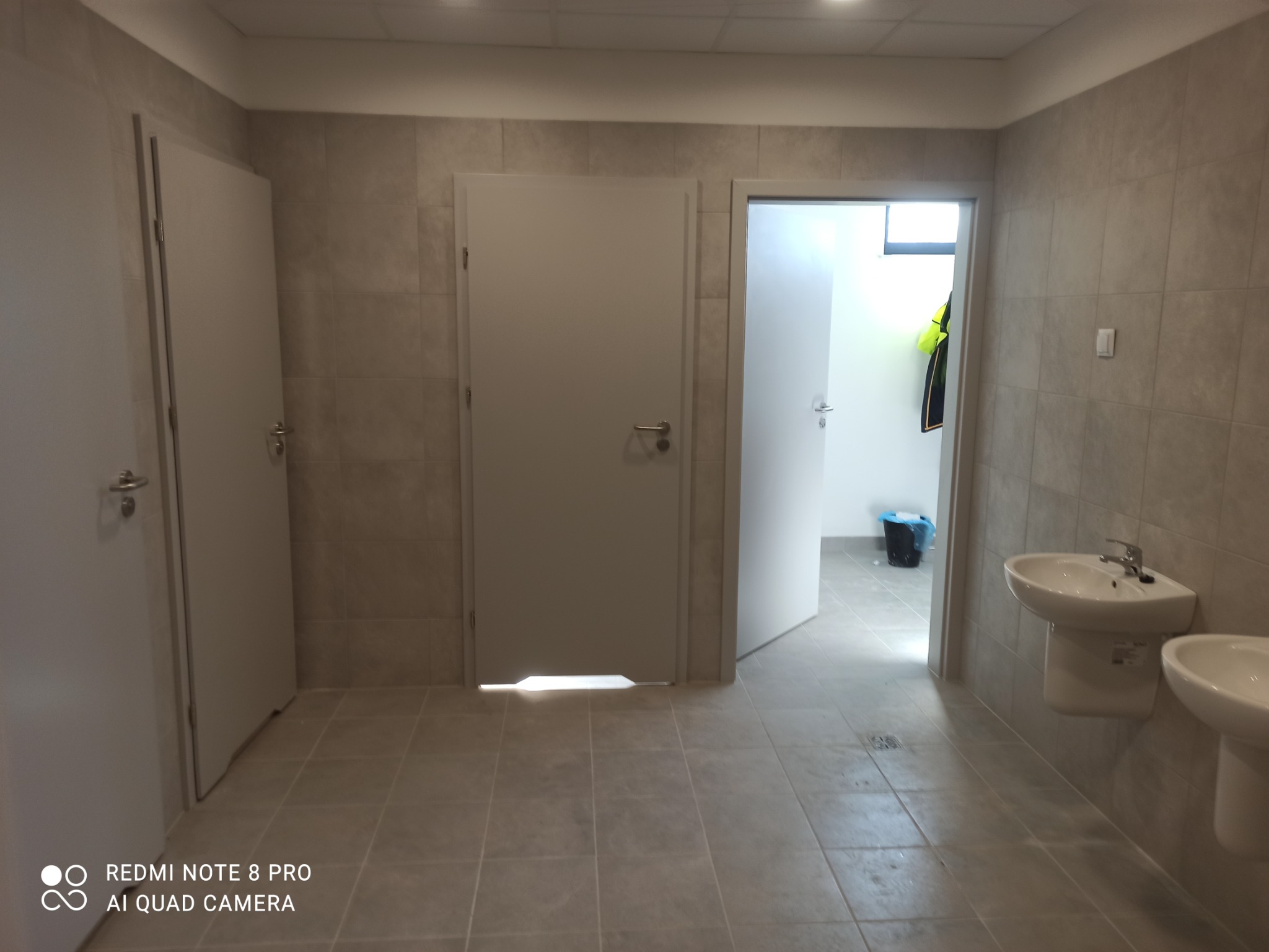 Wykończona łazienka z beżowymi płytkami, dwoma umywalkami i trzema białymi drzwiami, w tym jedne otwarte, z widocznym w tle workiem na śmieci i odblaskową kamizelką.